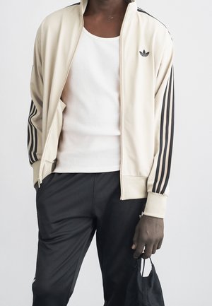 Homme portant une veste de survêtement Adidas crème et un pantalon noir, tenant un masque facial noir dans la main droite.