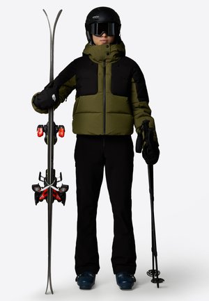Skioutfit med en grøn og sort jakke, sorte bukser, blå skistøvler og holder ski med røde bindinger samt skistave.