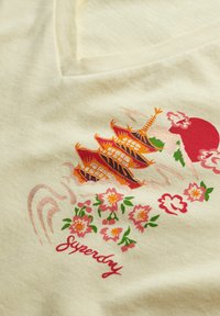 Lys gul t-shirt med et levende design af rød og orange pagode, pink blomster, og teksten "Superdry" i et afslappet skrifttype. Glat bomuldsstruktur.