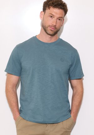 Mand med kort krøllet hår iført blå kortærmet T-shirt og beige bukser, stående med hænderne i lommerne mod en hvid baggrund.