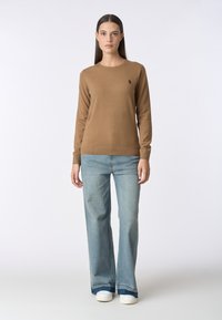 Maglione marrone a maniche lunghe realizzato in morbido tessuto, con scollatura rotonda e un sottile dettaglio del logo, abbinato a jeans azzurri a gamba larga e scarpe bianche.
