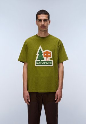Homme portant une chemise manches courtes vert olive avec logo arbre et cabane dans l’arbre et texte "NAPAPIJRI", debout devant un fond gris clair.