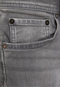 Primer plano de una tela de mezclilla gris que presenta un bolsillo delantero con costuras visibles, herrajes de metal y una sutil variación de textura.
