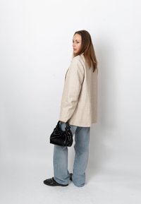 Femme aux longs cheveux portant un blazer beige et un jean bleu, tenant un sac à main noir, debout sur un fond blanc uni.