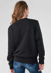 Sweatshirt noir à col rond, avec des poignets et un ourlet côtelés. Texture lisse, présentant une coupe décontractée et un design simple. Jeans visibles en dessous.