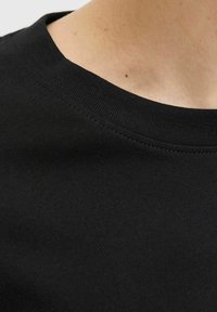 Primer plano del cuello y la parte superior del pecho de una persona que lleva una camiseta negra de cuello redondo con costuras visibles.