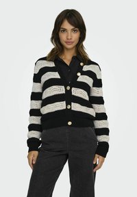 Cardigan tricoté à rayures noires et beiges avec un col en V, des fermetures à boutons et des poignets côtelés. Le tissu présente une texture à mailles ouvertes.