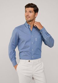 Lichtblauw blouse met lange mouwen en een zak aan de linkerkant, gecombineerd met crèmekleurige broek. Soepele stofstructuur.