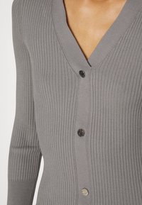 Gros plan d'une personne portant un cardigan gris côtelé cintré avec trois boutons visibles à motifs et un col en V.
