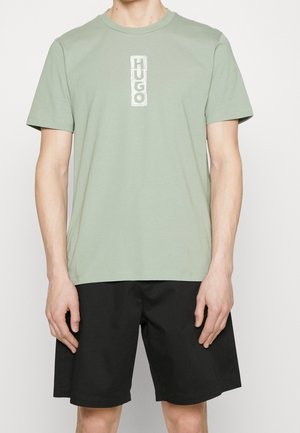 Homme portant un t-shirt vert sauge à manches courtes avec le texte vertical blanc "HUGO" et un short noir jusqu'aux genoux, debout devant un fond blanc.