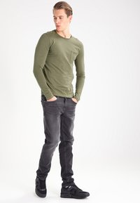 Olive green long-sleeve shirt with chest pocket, paired with dark gray jeans and black sneakers, showcasing a fitted design and smooth texture.
