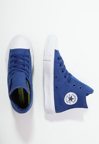 Blue high-top sneakers made of canvas, featuring white rubber soles, round toe caps, and a circular logo on the side. Laces are visible.