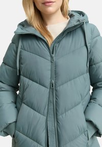 Pufferjack in zacht teal, met een rits aan de voorkant, een capuchon en een diagonaal gequilt patroon. De stof is glad met elastische mouwen.