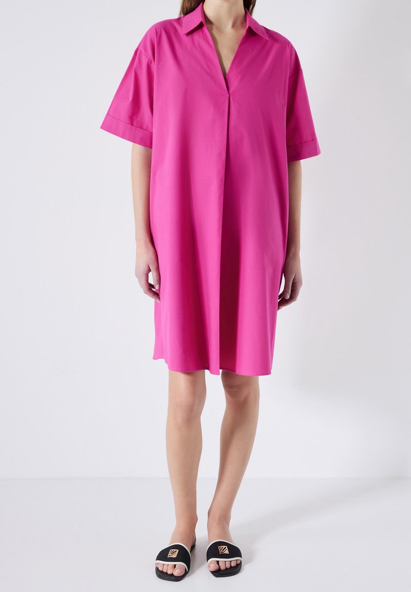 Femme portant une robe ample rose vif à longueur genou, avec manches courtes et col, assortie à des sandales noires sans bride.