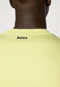 Sárga crewneck póló sima textúrával. Jellemzője egy fekete "BOSS" logó a hátsó részen, a nyakkivágás közelében. Egyszerű és modern dizájn.