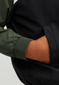 Crna jakna s zelenim manžetama, s elastičnim rebrastim detaljima. Glatka tekstura, s vidljivim džepom i detaljima šivanja. Pogled izbliza.