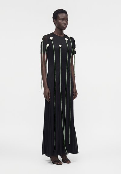 Ψηλή γυναίκα που φοράει μακρύ μαύρο φόρεμα με κοντά μανίκια, διακοσμημένο με κάθετα πράσινα στελέχη και λευκά λουλούδια μπροστά και στα μανίκια.