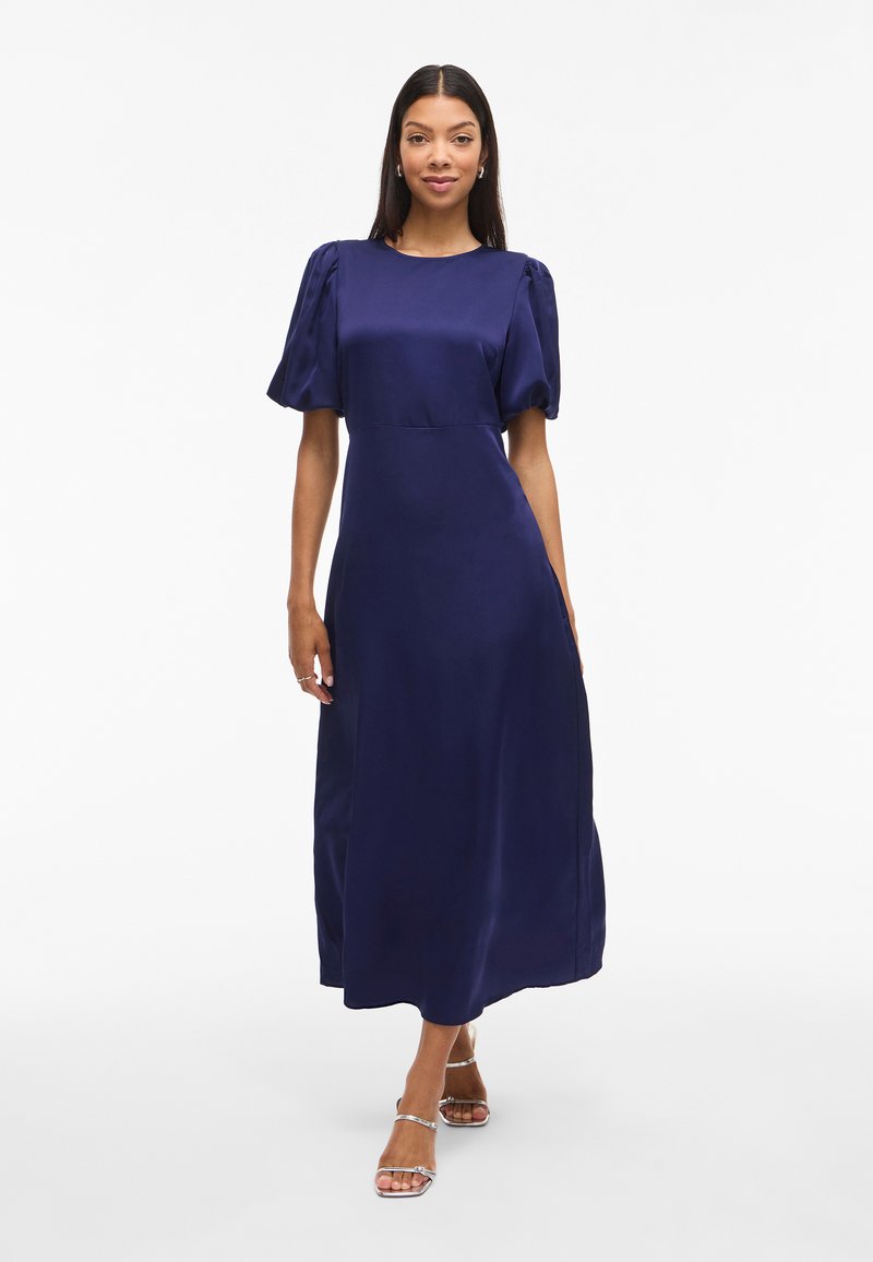 Žena v námořnické modré midi šaty s vykasanými rukávy a stříbrnými sandály na vysokém podpatku, stojící proti bílému pozadí.