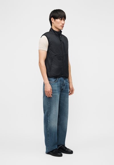 Νεαρός άνδρας που στέκεται πλαγίως, φοράει μαύρο γιλέκο με επένδυση, μπεζ κοντομάνικο πουκάμισο, φαρδιά μπλε τζιν και μαύρα αθλητικά παπούτσια σε απλό φόντο.