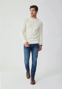 Man walking wearing white cable knit sweater, blue jeans, brown leather shoes, and a wristwatch on a plain light background.