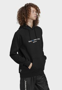 Sort hoodie med hætte og en frontlomme, prydet med hvid og blå tekst "Alle har en historie" og tre striber på ærmerne.
