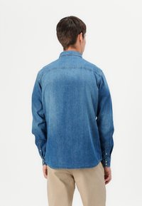 Camicia di jeans azzurro chiaro con maniche lunghe, colletto e polsini a bottone. Presenta una texture sottile e un orlo curvo, indossata sopra pantaloni beige.