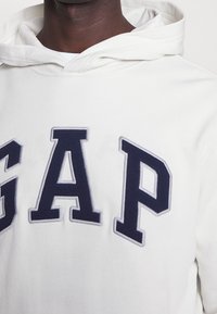 Felpa bianca con tasca a marsupio frontale, con grandi lettere ricamate in blu navy "GAP" in stile blocco. Tessuto morbido con cappuccio.