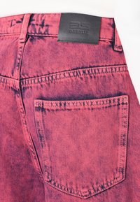 Jean en denim rose avec une texture usée, comportant une poche arrière et un écusson en cuir noir au niveau de la taille.