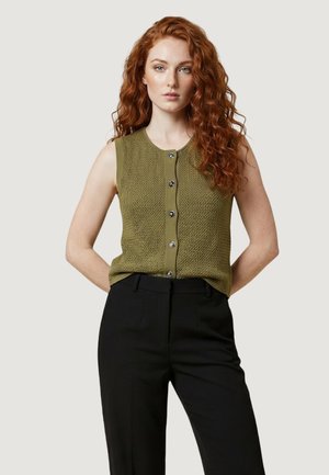 Donna con lunghi capelli rossi ricci che indossa un gilet senza maniche verde oliva lavorato a maglia con bottoni e pantaloni neri a vita alta.