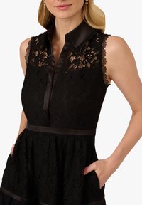 Abito nero in pizzo con colletto in satin, design senza maniche, bordi in pizzo ondulati e vita cintrata con tasche.