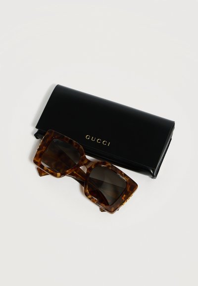 Τετράγωνα γυαλιά ηλίου με φιλοτεχνία σε σχήμα χελώνας και σκουρόχρωμους φακούς δίπλα σε μαύρη θήκη Gucci με χρυσά γράμματα σε λευκό φόντο.