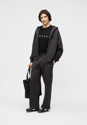 Νεαρό άτομο ντυμένο με μαύρη μπλούζα με κουκούλα, μαύρο γραφικό μπλουζάκι, φαρδύ σκούρο παντελόνι φόρμας, μαύρα αθλητικά παπούτσια και κρατώντας μαύρη υφασμάτινη τσάντα, στέκεται μπροστά σε γκρι φόντο.