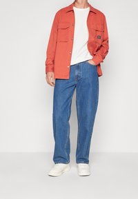 Röd bomullsskjorta med två bröstfickor, matchad med lösa blå denimjeans och vita sneakers. Enkel vit t-shirt under.