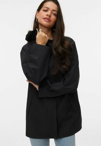 Parka imperméable noire avec capuche, fermeture à boutons sur le devant, poches latérales et coupe décontractée. Texture lisse, design minimaliste.