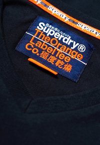 Zwarte T-shirt stof met een V-hals, voorzien van een blauwe patcheslabel met witte en oranje tekst, en een oranje accent langs de halslijn.