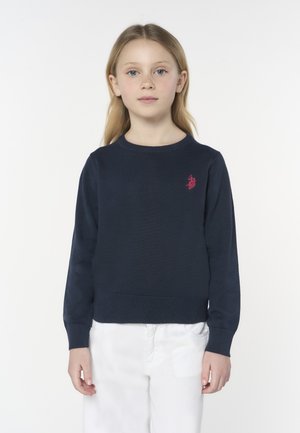 Ragazza giovane con capelli biondi lunghi che indossa un maglione blu navy con logo rosa e pantaloni bianchi, in piedi davanti a uno sfondo bianco uniforme.