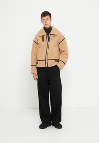 Beige fleecejakke med lynlås, brune accenter og krave; båret med sorte vide bukser og sorte sko.