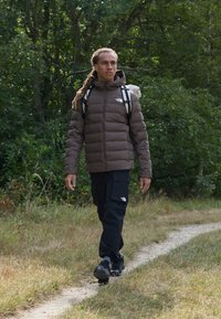 Brown puffer jacket with a hood, black cargo pants, and black footwear. The person carries a light backpack and walks on a dirt path in greenery.