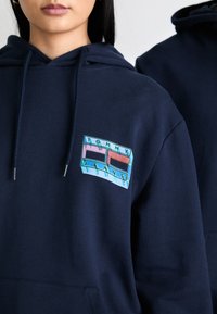 Sweat à capuche bleu marine en mélange de coton, avec un patch logo brodé multicolore comportant le texte "TOMMY JEANS". Texture douce et coupe décontractée.