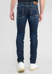 Jeans de mezclilla ajustados de color azul oscuro con un acabado texturizado, diseño de cinco bolsillos y detalles de costura discretos. Llevados con zapatillas deportivas blancas.