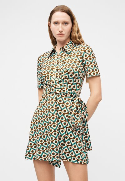 Γυναίκα που φοράει κοντομάνικο φόρεμα με γιακά, με γεωμετρικό σχέδιο σε καφέ, τιρκουάζ και άσπρο, δεμένο στη μέση με φιόγκο.