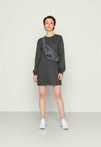 Mulher jovem está de pé, vestindo um vestido longo de manga escura, uma bolsa crossbody cinza da Nike e ténis brancos, em frente a um fundo claro e liso.