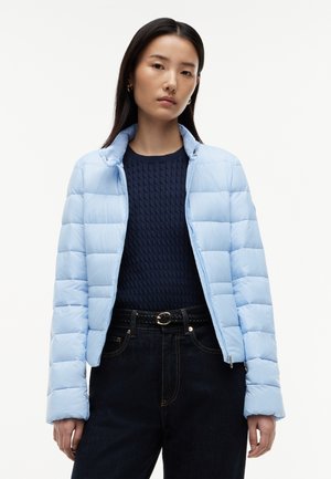 Ung kvinde iført en lyseblå quiltet jakke over en marineblå kabelstrikket sweater med mørke jeans og et flettet sort bælte, stående neutralt.