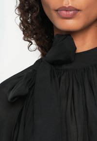 Black blouse featuring a tie neck, pleated design, and lightweight fabric. Close-up view highlights texture and draping details.