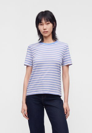 Person står mod enkel baggrund iført en kortærmet blå og hvidstribet t-shirt og mørkeblå bukser, hænderne afslappet langs siden.