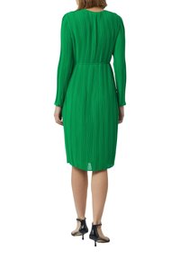 Robe plissée verte à manches longues, taille cintrée et ourlet au genou. Associée à des talons ouverts noirs.