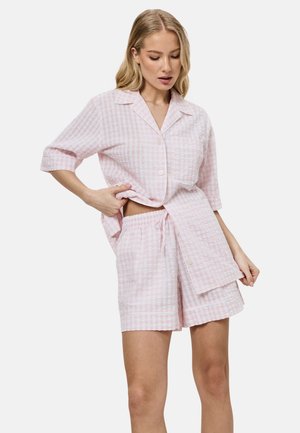 Vrouw draagt een loszittend, roze-wit geruit overhemd met korte mouwen en knopen en bijpassende shorts met elastische taille, staand tegen een witte achtergrond.