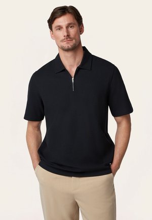 Man draagt een zwarte korte mouw polo met een rits aan de voorkant en beige broek, staat met handen in de zakken tegen een effen achtergrond.