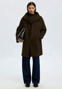 Mulher vestindo um casaco castanho-escuro, calças largas azuis-marinho, sapatos pretos e segurando uma bolsa preta contra um fundo simples.