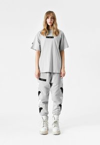 Camiseta gris de gran tamaño con una etiqueta negra, combinada con joggers grises que presentan patrones de alas en blanco y negro, acompañados de botas blancas.